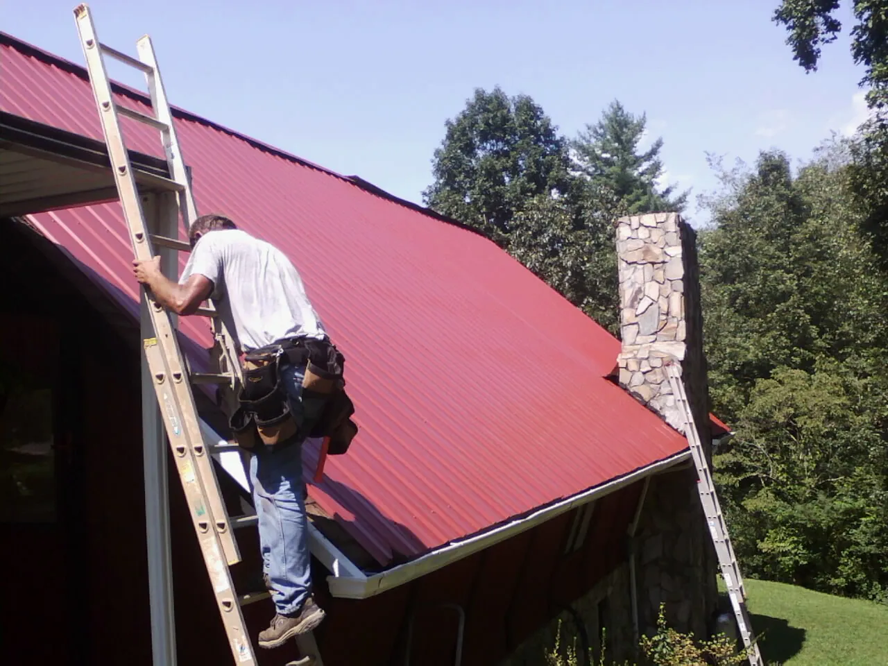 Roofer on a ladder beside a red metal roof and stone chimney — Soffit & Fascia Repair by Roofing Brothers in Bishop